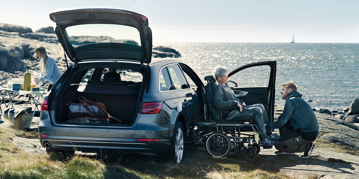 Car with its boot open by the coast while a family helps an older person in a wheelchair