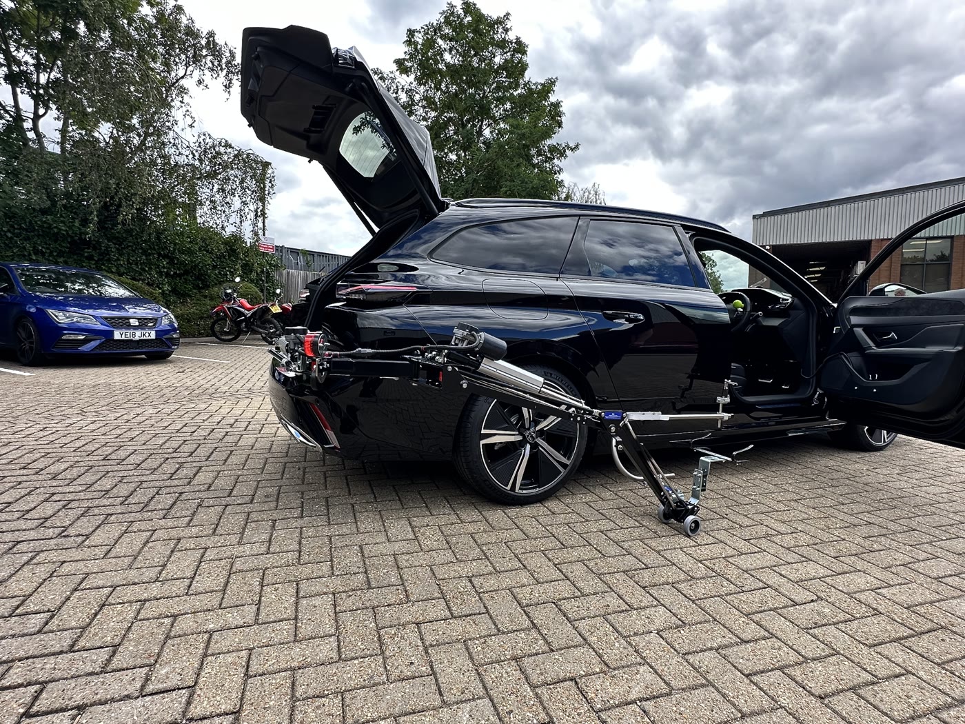 Abi Loader arm reaching from the boot to the open driver's door of a black estate car